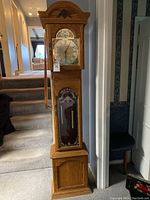 Full view of tall wooden grandfather clock with glass door revealing pendulum and weights, standing in a hallway.