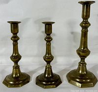 Three brass candlesticks: one taller single and two shorter identical ones, standing upright against a white background.