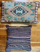 Two vintage decorative throw pillows placed side by side on wood floor. The top pillow is rectangular with an Aztec geometric woven pattern in brown, blue, green, and beige. The bottom pillow is square with horizontal Kilim style stripes in blue, pink, and white.