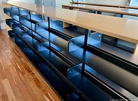 Long shot showing multiple black metal shelving units joined in a row topped with wooden boards, standing on wood floor.
