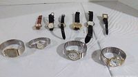Overview of 10 assorted wristwatches arranged on a white surface. Watches have different strap types including metal expandable, leather, and synthetic bands. Watch faces are analog with various round and square dial shapes.