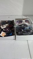Two boxes containing multiple pairs of sunglasses in various styles and colors, some with packaging and tags