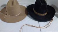Two cowboy hats side by side on a white surface. Left is tan Canyon Trails Range Rider with discoloration and chin strap. Right is black Lanning hat with decorative band and chin strap, both showing wear.
