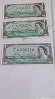 Three Canadian one dollar bills showing front side with Queen Elizabeth II portrait, two bills 1967 and one bill 1954. Bills show wear, creases, but no tears.