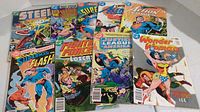 Photo showing nine DC Comics including Steel, Justice Society, Superman, Flash, Fightin' Force, Justice League of America, and Wonder Woman comics, colorful covers with slight wear.