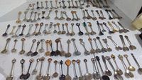 Wide shot showing the entire collection of about 103 souvenir spoons laid out on a white background in an organized grid, displaying varied designs and decorative handles.