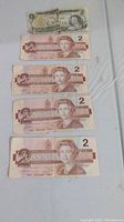 All five Canadian bills laid out visible the $1 bill on top and four $2 bills below it showing wear and handling marks.