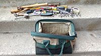 Photo showing tool bag partially open with various hand tools laid out above it including wooden mallet, screwdrivers, sockets, pliers, and other tools.