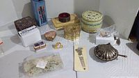 Photo showing multiple trinket boxes and decorative items arranged on a white table, including round ivory-colored box, gold-tone heart box, shell-shaped silver-tone item, small decorative rectangular boxes, and beaded bracelet.