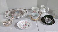 Eight fine china pieces including assorted creamers, sugar bowls, teacup with saucer, trinket dish, and platter shown on white surface.