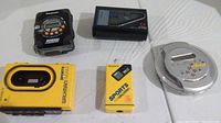 Photo showing all 5 devices: black Panasonic Walkman, black cassette player with AM/FM radio, yellow Sony Sports Walkman with missing front cassette window, yellow Sony Sports FM Walkman tuner, silver portable CD player.