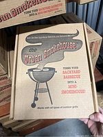 Single Urban Smokehouse kit box held in hand, showing front graphic and text explaining it turns backyard BBQ into a mini-smokehouse.