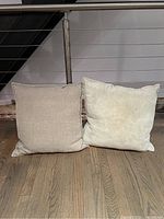 Two Arhaus throw pillows side by side on a wooden floor against a metal and cable railing. One pillow is beige woven fabric, the other is light cream soft fabric.