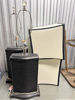 Pair of black woven-pattern table lamps with white and black-trimmed rectangular lampshades on top. Both lamps have power cords attached.