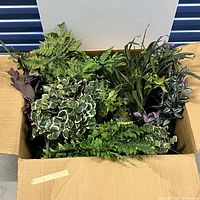 Box containing assorted faux greenery plants with various leaf shapes and colors.