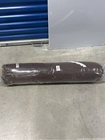 Brown fabric hammock tightly rolled and sealed in plastic packaging, placed on a concrete floor.