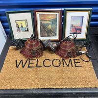 Photo showing three framed art prints arranged behind two fabric pendant lamps atop a natural coir doormat with 'WELCOME' in black letters.