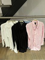 Five Jos A Banks men's dress shirts displayed on hangers including three white, one black, and one pink shirt.