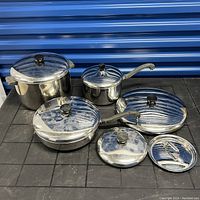 Full set of six stainless steel pots and pans with lids displayed on black table against blue background