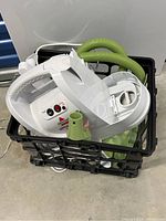 Top view of basket containing the white main steamer unit with power and heat indicators, green hose, and some attachments.