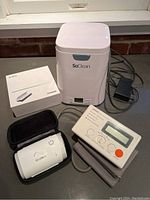 Lot of medical devices on grey countertop, including SoClean CPAP cleaner, Omron pulse oximeter, Phonak D-Dry box, and ResMed device in black case