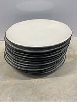 Stack of 10 white stoneware plates with thin black rim, showing the side profile and top plate surface.