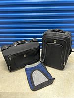 Two black Travelpro soft-sided suitcases and one blue shoe bag arranged side by side against a blue metal door.