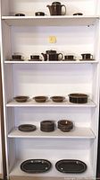 Full white shelving unit with all pottery items arranged by type on shelves highlighting mugs, bowls, plates, and servingware
