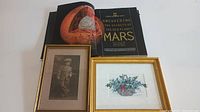 National Geographic book open to a detailed illustration of Mars, alongside framed vintage photo of young boy in early 1900s attire and signed framed painting of holly berry basket.