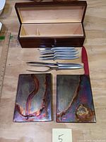Full lot laid out on wood table: wooden box open, six carving knives, carving fork, sharpening steel, two rectangular artisan metal art pieces.