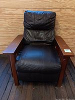 Front view of the large mission style armchair with black cushioned seat and backrest made of leatherette, dark wooden armrests with slats on sides, in used condition with visible creases.