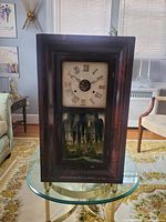 Front view of antique wooden wall clock showing rectangular dark wood frame, clock face with Roman numerals and glass door with painted lower panel