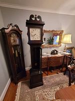 Front view of the tall case clock showing dark wood case, white clock face with visible damage, decorative top, and pendulum visible in glass panel.