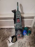 Front view of Bissell Pro 2X carpet cleaner with attachments, solution bottles, instruction manual, and mesh accessory bag on floor.