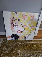 Front view of a large square canvas print with abstract art including stylized human faces and figures in mixed colors including cream, yellow, black, and pink.