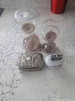 Overview of all four items: two hourglasses, embossed pill box, and floral lighter on a countertop.