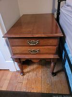 Front view of brown wooden end table with two drawers, brass handles, and turned front legs on hardwood floor next to bed.
