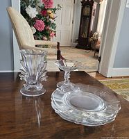 Clear glass vase, pedestal dessert dish, and one plate on wooden table with chair and flowers in background.