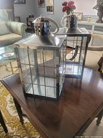 Pair of metal lanterns on wooden table in living room; both silver-tone with glass panes and rope handles.