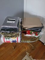 Stack of assorted linens including folded quilts, solid color fabrics, and patterned pieces on glass table near a corner wall.