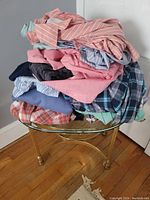 Pile of men's shirts on a glass and brass table showing variety of colors and patterns including stripes, plaid, solid colors.