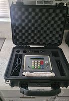 Control device inside hard plastic case with foam padding. Device features numeric keypad, intensity knob, and wired accessory cables.