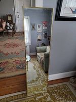 Full-length rectangular wall mirror with a gold frame standing on the floor reflecting room with floral rug, armchairs, and framed art.