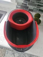 View of black and red O-Cedar spin bucket showing spin basket and handle on white countertop.