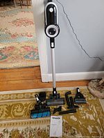 Image showing the entire cordless stick vacuum upright along with multiple cleaning attachments and user manual on floor.