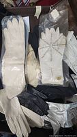 Gloves in various colors including white, black, beige, and silver. Different textures and some gloves have button accents. Mixture of leather and fabric.