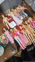 Photo showing full lot of 15 dolls closely arranged on wooden floor with some dressed and some nude. Includes Barbie, Ken, Jo-Jo, Ariel dolls.
