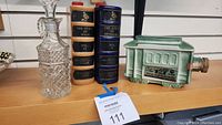 Four items displayed on shelf: two ceramic book decanters, one clear cut glass bottle with stopper, green trolley car decanter