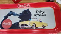 Coca-Cola metal tray featuring vintage 1950s advertisement with person drinking Coca-Cola and yellow convertible, 'Drive refreshed' text visible.