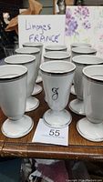 All twelve white Limoges coffee cups with silver trim and monogrammed letter 'J' displayed on a wooden table, showing their shape, size, and markings.